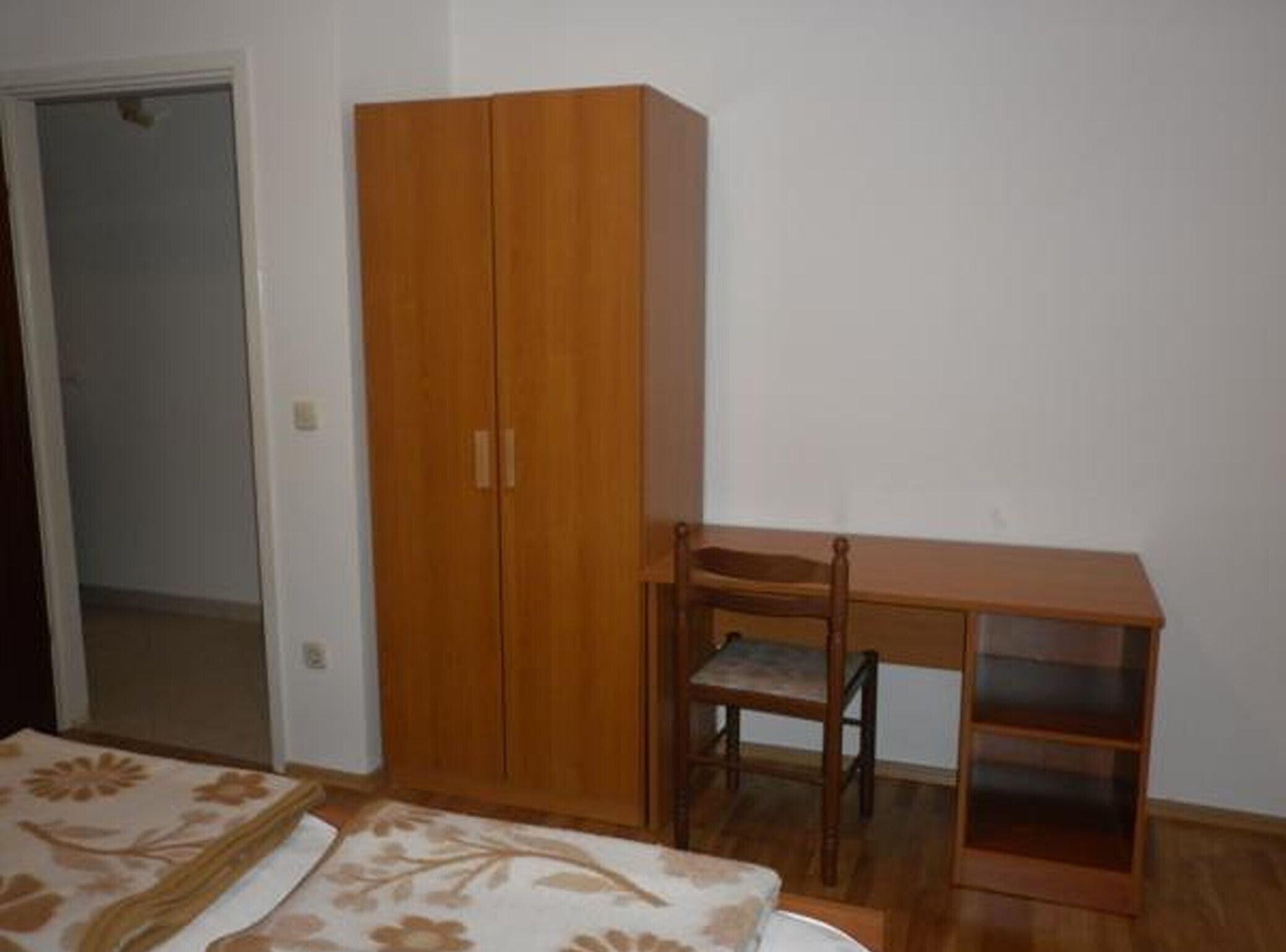 A bedroom with a wooden wardrobe, a wooden desk with a chair, and a bed with floral bedding.