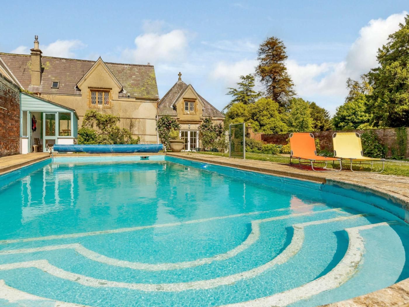 A swimming pool with clear blue water, surrounded by a brick wall and a house with a pitched roof.