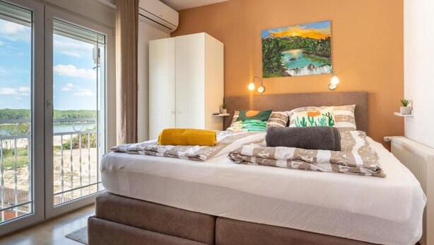A bedroom with a large bed, a painting, a nightstand, and a view of a body of water through large windows.