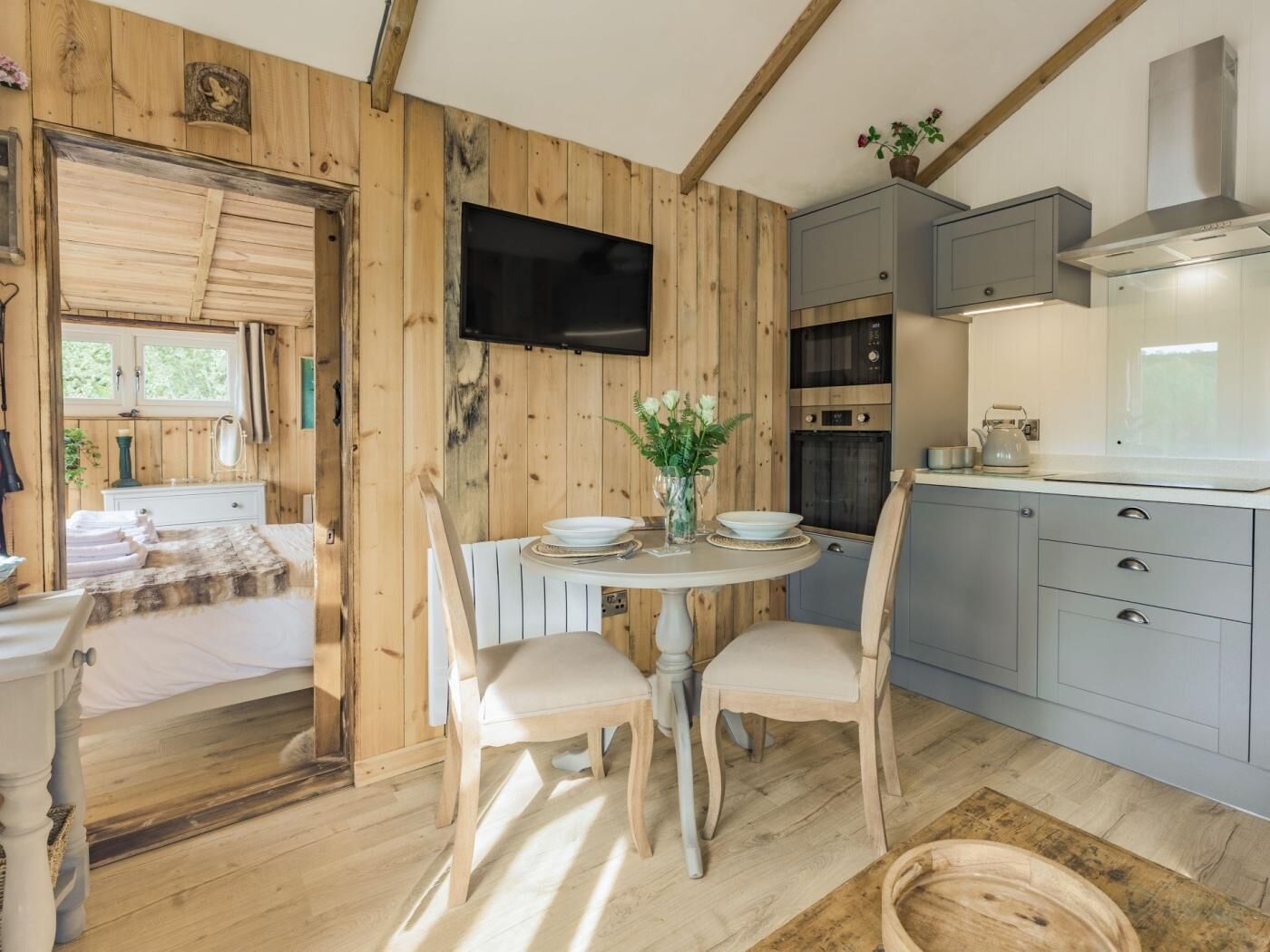 A cozy cabin interior with a wooden interior, a dining area with chairs and a table, a kitchen with cabinets and a stove, and a television mounted on the wall.