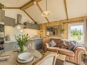 A cozy living room with a brown leather sofa, a dining table set for two, and a kitchen with wooden paneling and modern appliances.