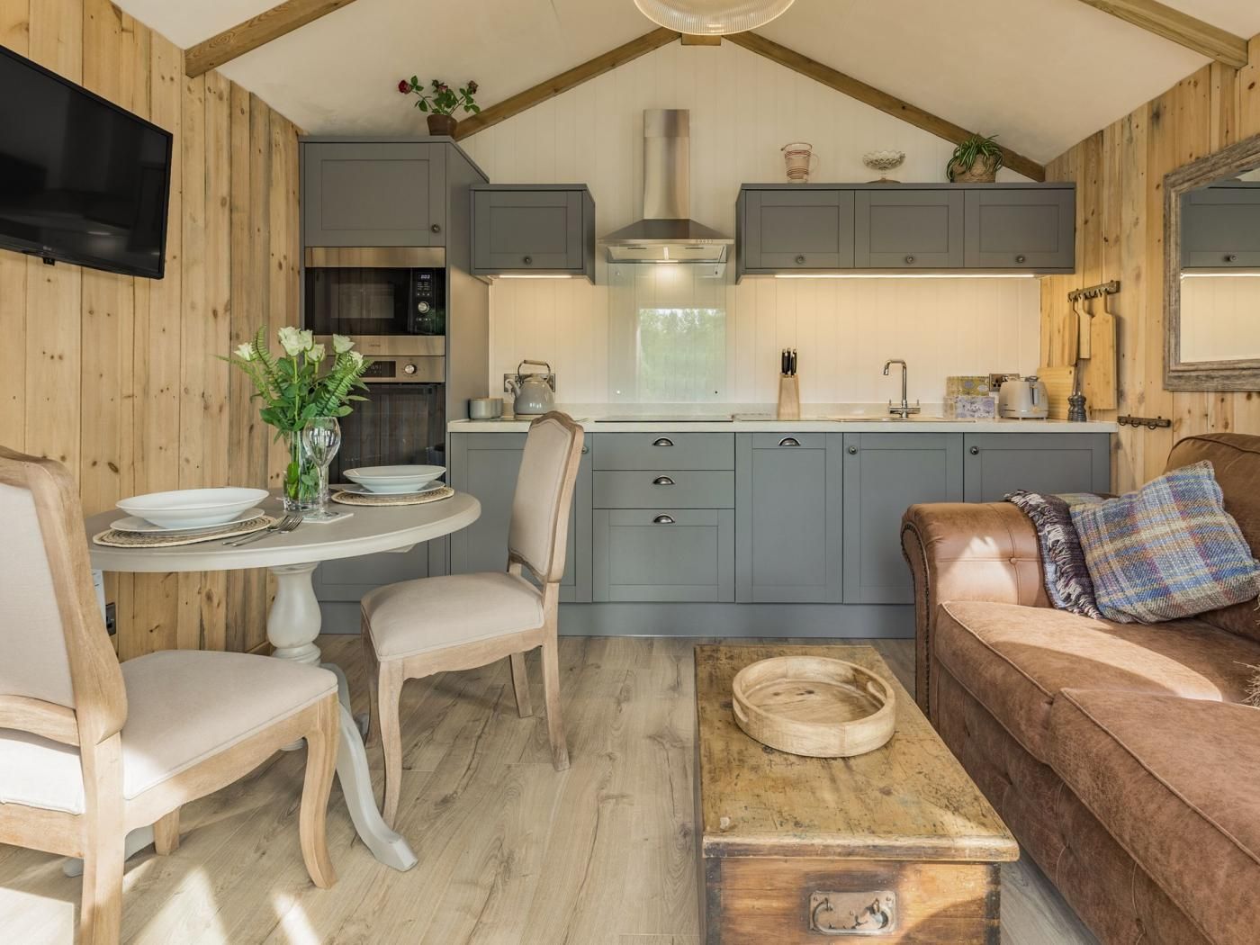 A cozy kitchen with grey cabinets, a wooden dining table with chairs, and a leather sofa with a plaid pillow.
