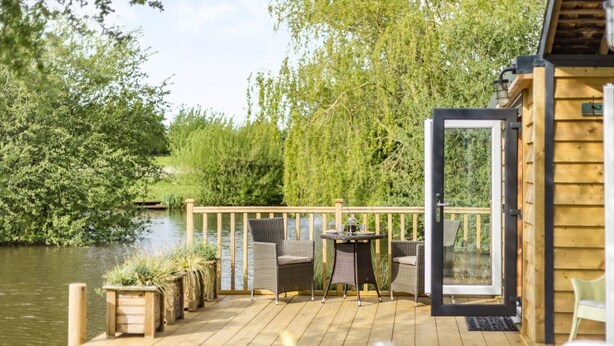Une terrasse en bois, agréée de meubles d’extérieur, d’une table et de chaises, qui offre une vue sur une étendue d’eau et est entourée d’arbres.