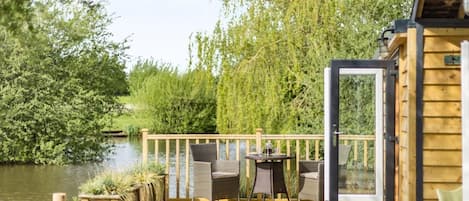 Eine Holzterrasse mit Gartenmöbeln, einem Tisch und Stühlen mit Blick auf ein Gewässer, umgeben von Bäumen.