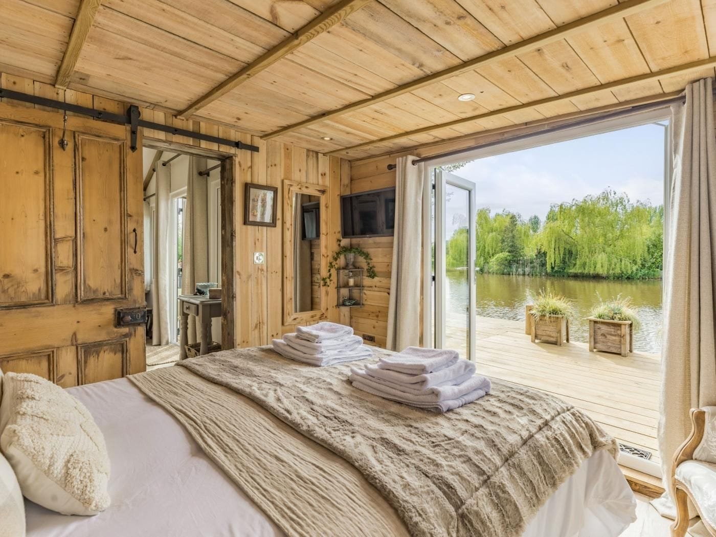 A wooden cabin bedroom with a bed, towels, a mirror, and a view of a river.