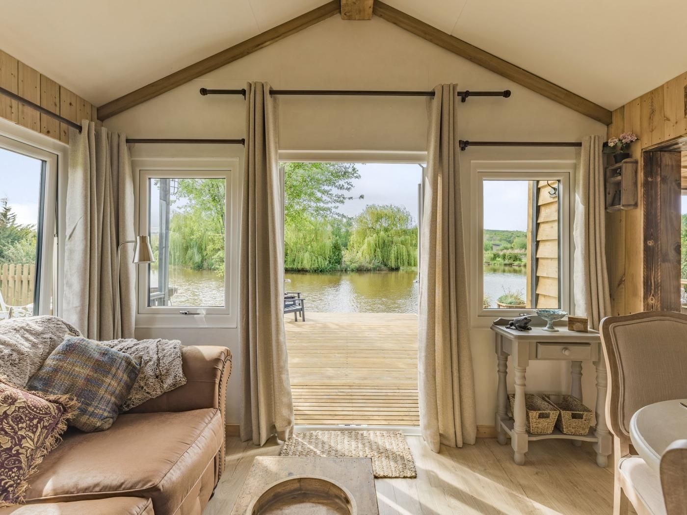 A cozy room with a wooden ceiling, a sofa with a throw blanket, a small table with a lamp, and a view of a river through large windows.