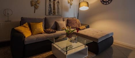 A living room with a sofa, ottoman, glass coffee table, wall art, and a clock.