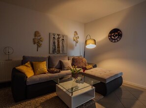 A living room with a sofa, ottoman, glass coffee table, wall art, and a clock. - Classic One Bedroom Apartment with Terrace (Vukovar)