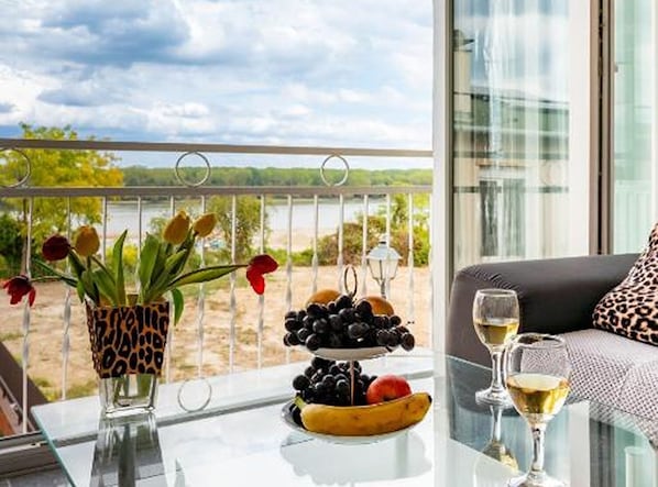 Un balcón con una mesa, un florero con flores, un frutero y una copa de vino.