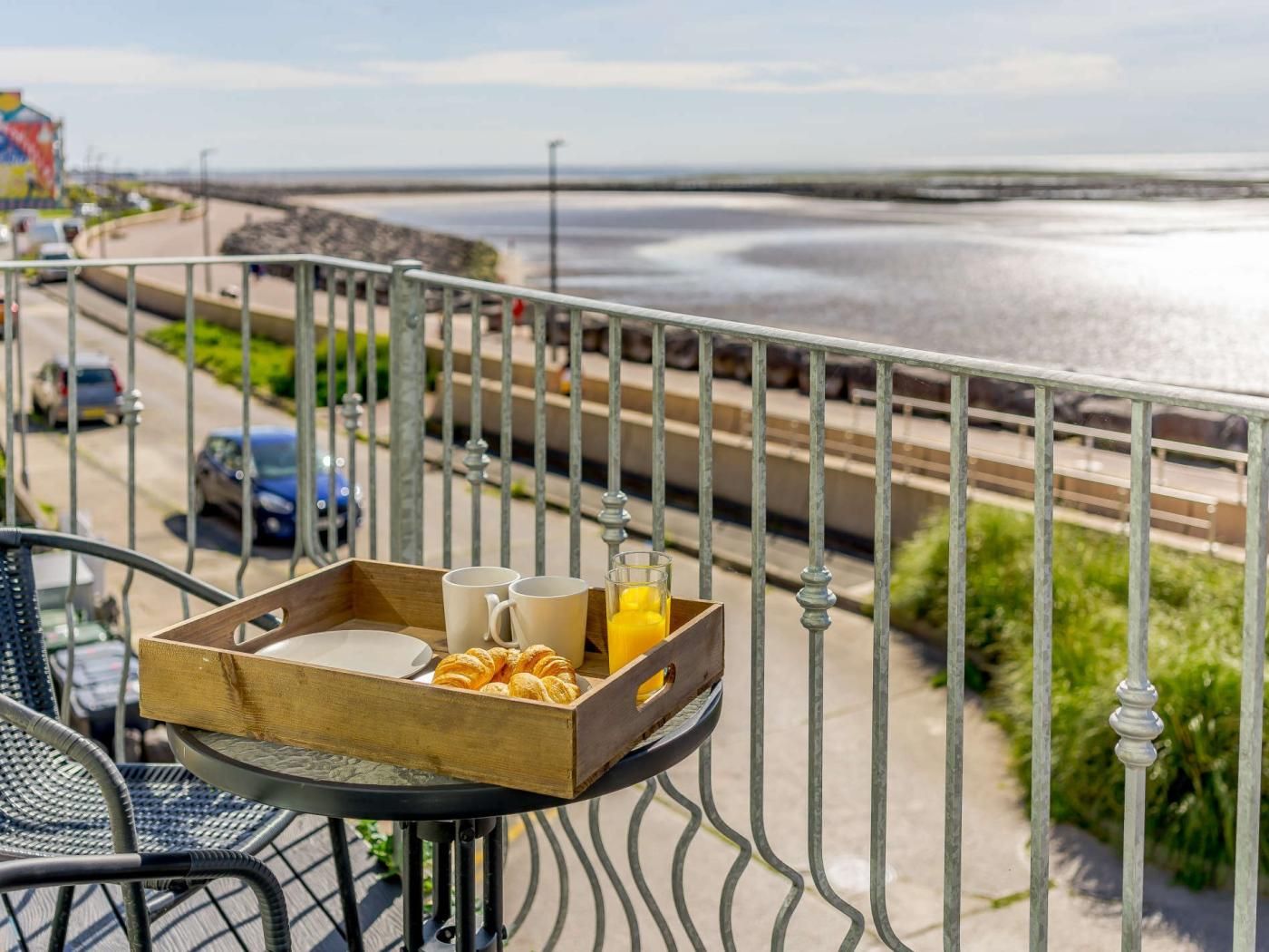 A balcony with a table set for breakfast, overlooking a beach and a road.