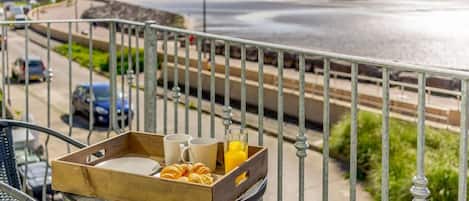 A balcony with a table set for breakfast, overlooking a beach and a road.