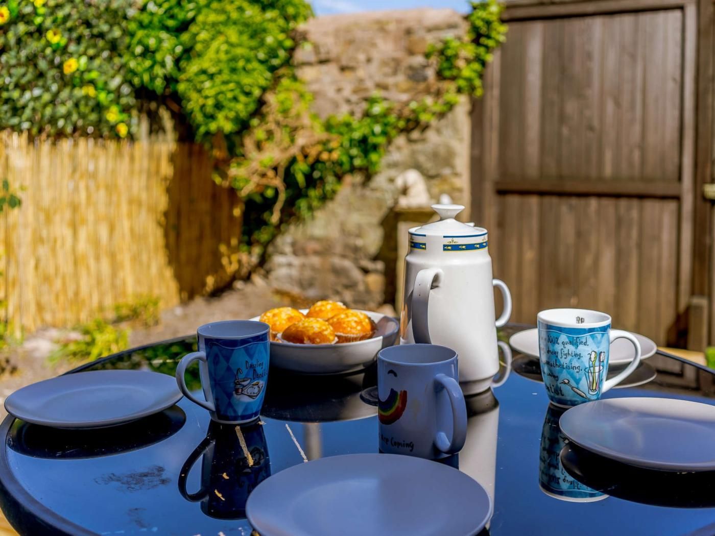 A table set outdoors with a coffee pot, a plate of muffins, and two mugs.