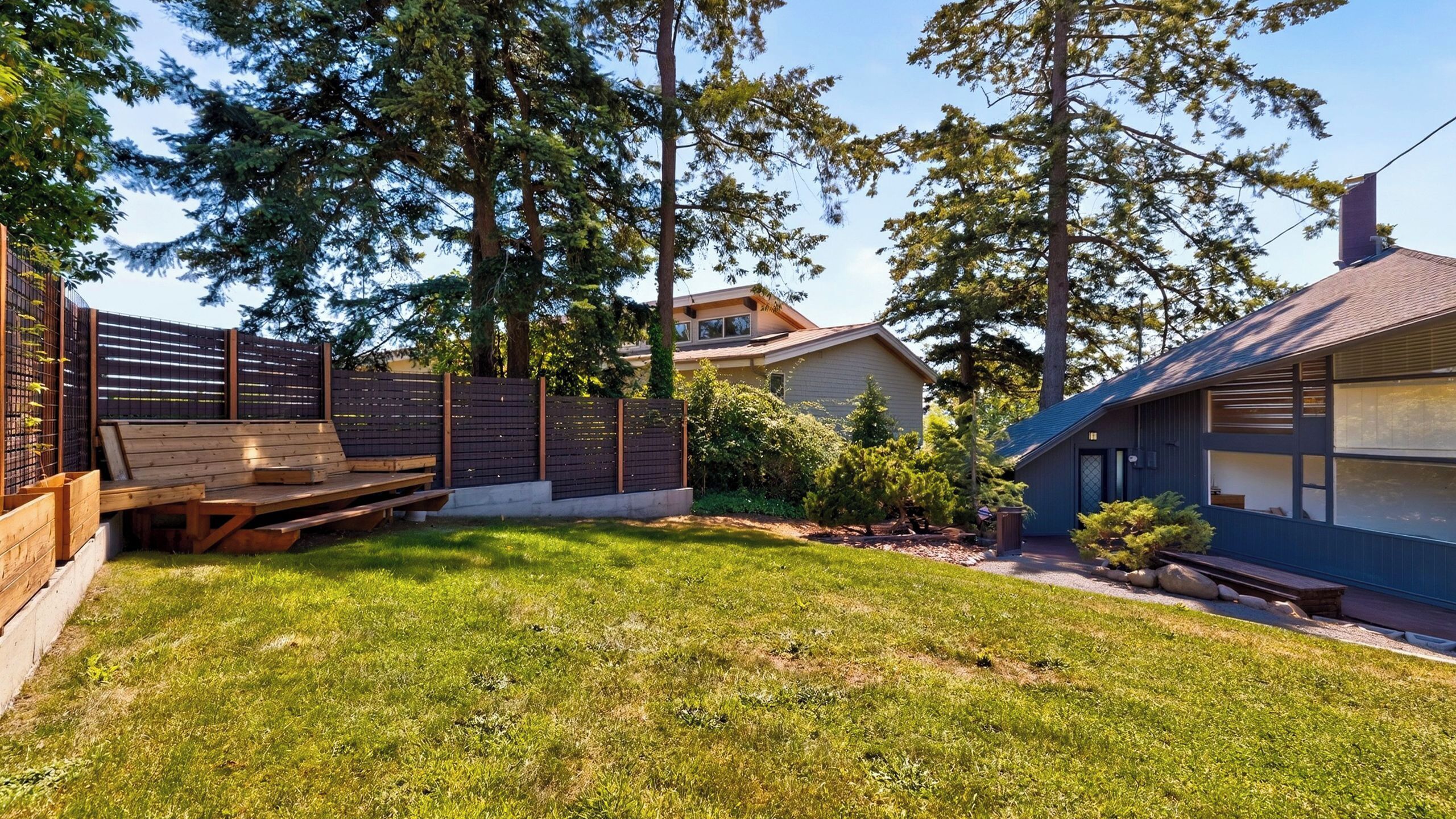 Ein Garten mit einer Holzterrasse, einem kleinen Gartenhaus und einem Haus mit dunkler Fassade.