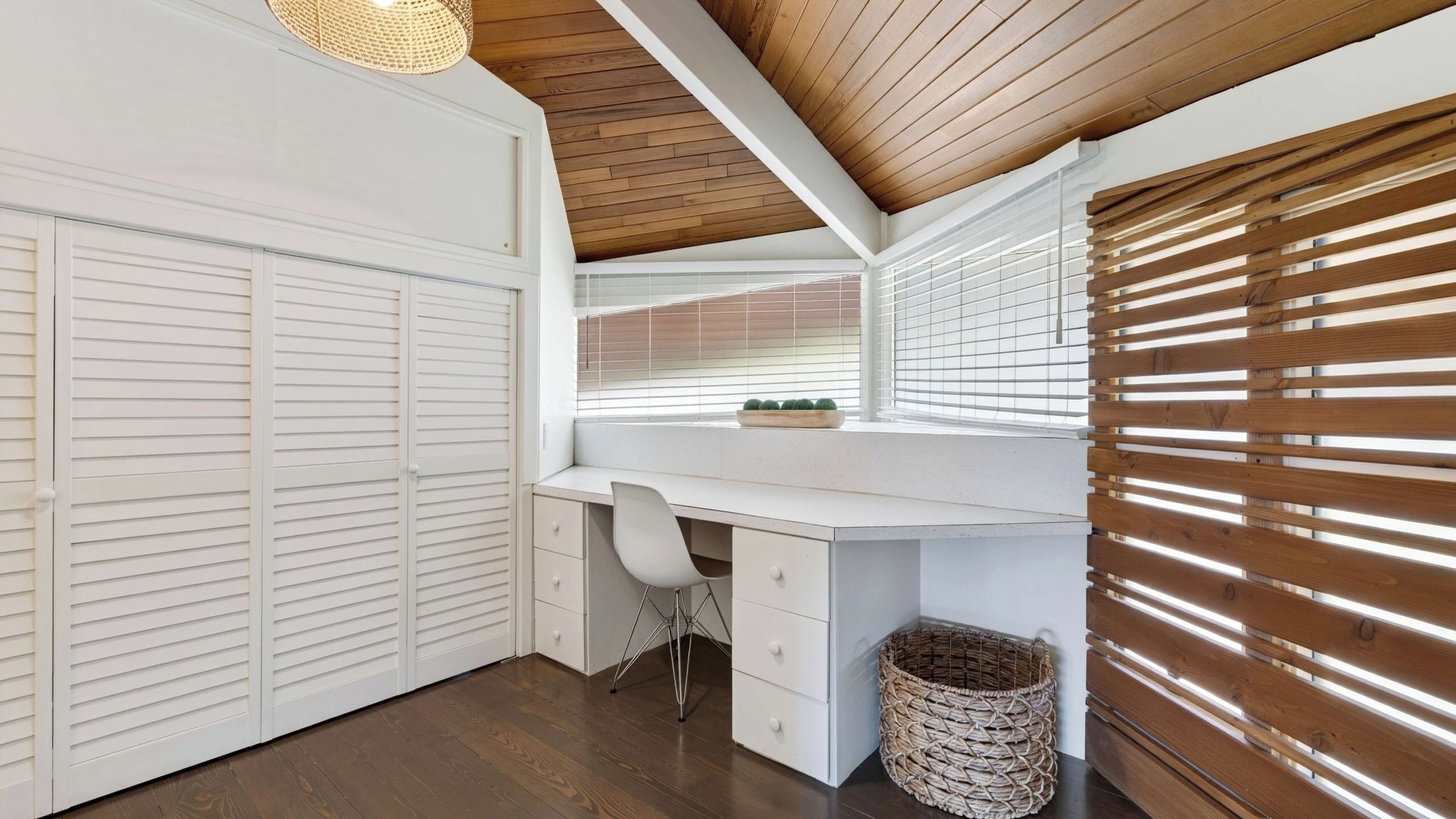 A room with a white desk, a chair, a basket, and a window with blinds.