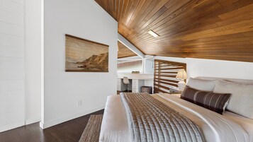 A bedroom with a wooden ceiling, a bed with a quilt, a desk, and a painting on the wall.