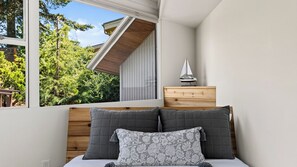 A bedroom with a bed, a nightstand, a window with a view of greenery, and a sailboat model. - The Grove at Mutiny Bay by Avantstay Walk to Beach Shared Pool + Pickleball Court + Water Views (Freeland)