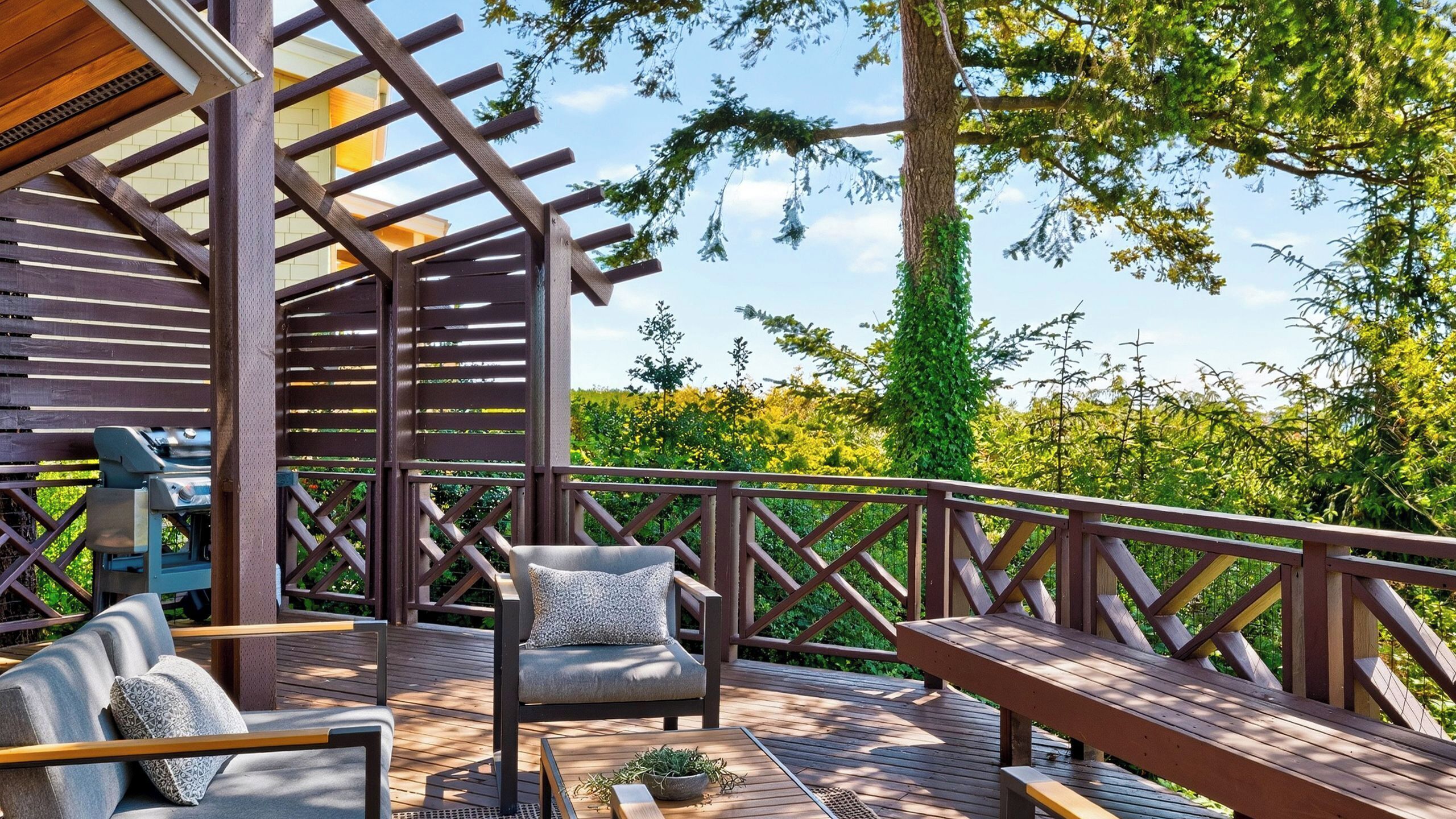 Una terraza de madera con una pérgola, mobiliario exterior y vistas a follaje.