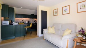 Ein Wohnzimmer mit einer Couch, einem kleinen Tisch mit einer Vase und einer Küchenzeile mit grünen Schränken und einem Esstischstuhl.