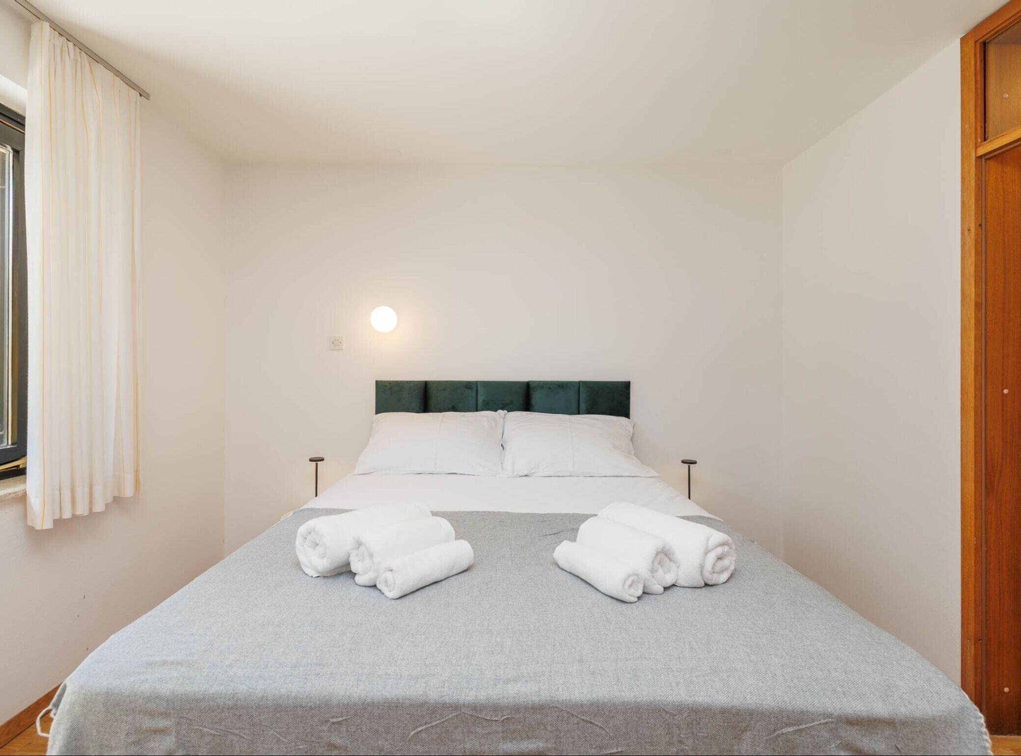 Una habitación de hotel con cama, dos almohadas, mesita de noche, lámpara y ventana con cortinas.
