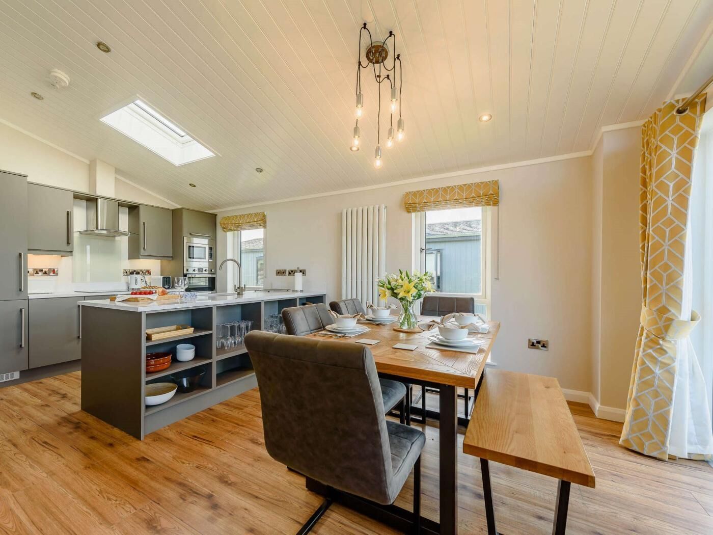 A modern kitchen with a central island, a dining area with a wooden table and chairs, and a large window with curtains.