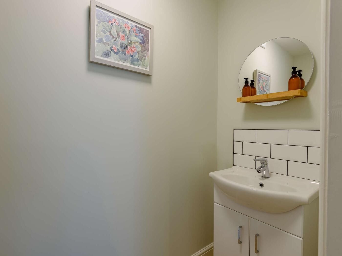 Un baño con un lavamanos blanco, un espejo, una obra de arte floral enmarcada y un estante con artículos de aseo.