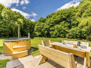A wooden hot tub with steps, a picnic table with benches, and a grassy area with trees in the background.