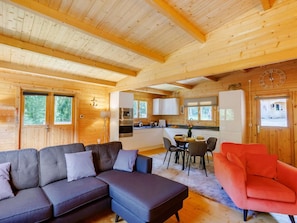 Ein Ferienhaus-Interieur mit einer Couch, einem roten Sessel, einem Esstisch mit Stühlen, einer Küchenzeile und einer Wanduhr.