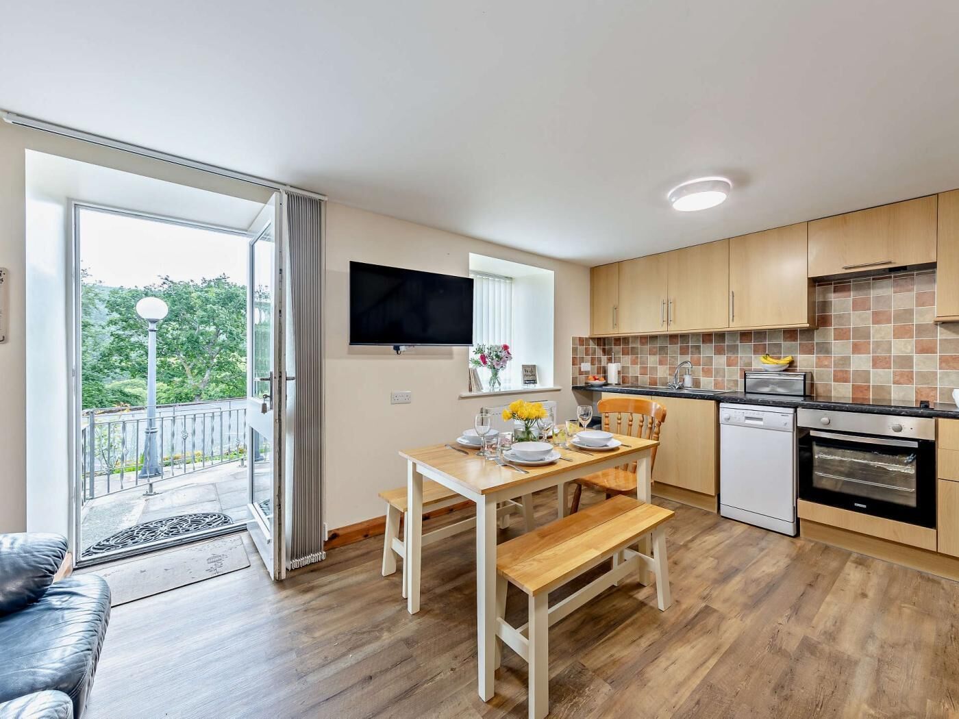 Eine moderne Küche mit Holzschränken, einem Esstisch mit Bänken, einem eingebauten Backofen und einem Fernseher, der über der Arbeitsplatte angebracht ist.