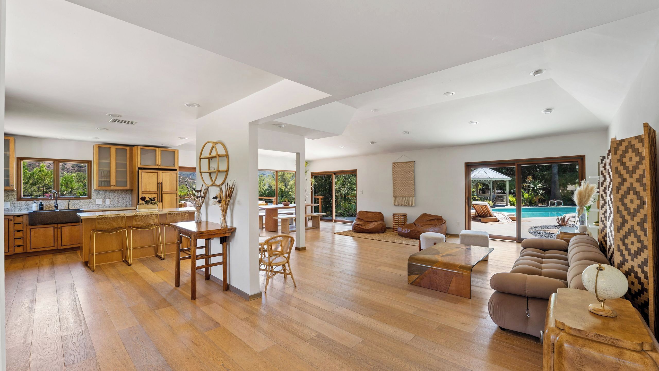 A spacious living area with a wooden floor, a dining table, sofas, and a large window overlooking a pool.