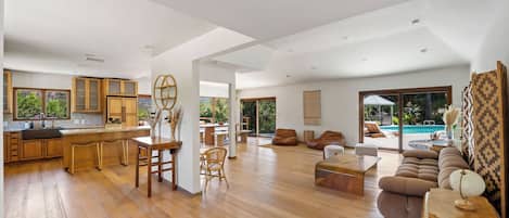 A spacious living area with a wooden floor, a dining table, sofas, and a large window overlooking a pool.