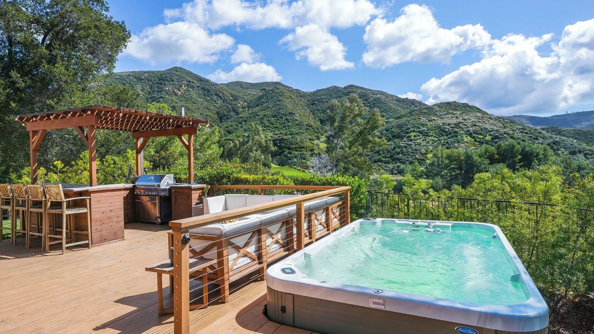 A wooden deck with a hot tub, outdoor seating, and a barbecue area overlooking a mountainous landscape.