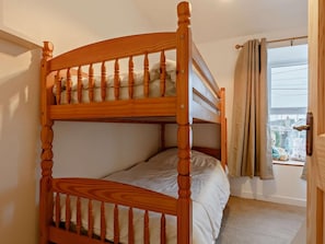 Uma cama de beliche com estrutura de madeira e roupa de cama branca, uma janela com cortinas e vista para prédios lá fora.