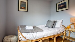 Uma cama de solteiro com cobertor e almofadas cinzas, uma cadeira de vime, uma pequena mesa de cabeceira com abajur, e duas molduras com quadros na parede.