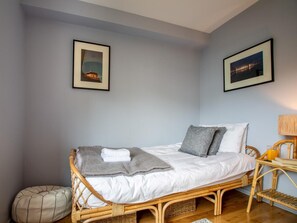 Ein Einzelbett mit grauer Decke und Kissen, ein Korbstuhl, ein kleiner Nachttisch mit Lampe und zwei gerahmte Bilder an der Wand.
