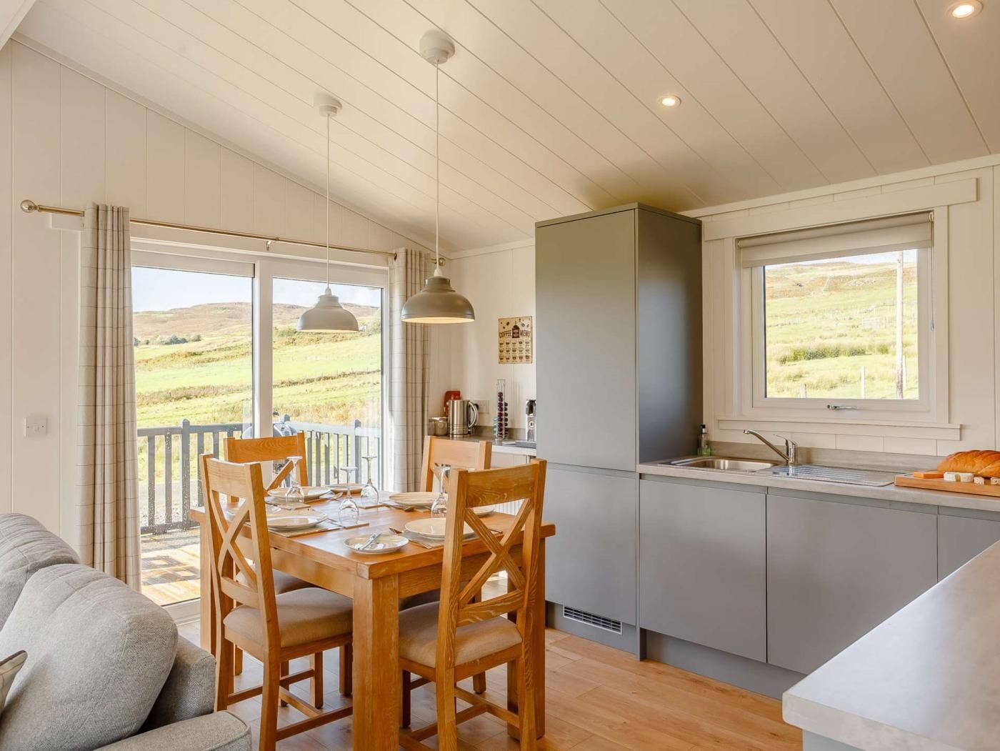 A dining area with a wooden table and chairs, a kitchen with a window, and a view of the countryside.
