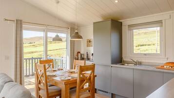 A dining area with a wooden table and chairs, a kitchen with a window, and a view of the countryside.