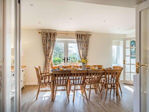 A dining area with a wooden table and chairs, a window with curtains, and a view of greenery outside. - 5 Bed in Norwich (oc-87444) (Southrepps)