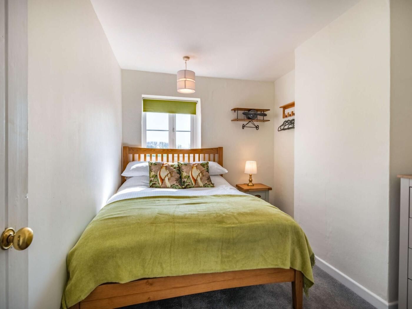 A bedroom with a wooden bed, a green bedspread, a bedside lamp, a window with a green blind, and a wall-mounted airplane decoration.