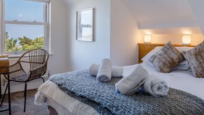 A bedroom with a bed, two bedside lamps, a chair, a window with a view, and a framed picture on the wall. - 3 Bed in Woolacombe (oc-87573) (Mortehoe)