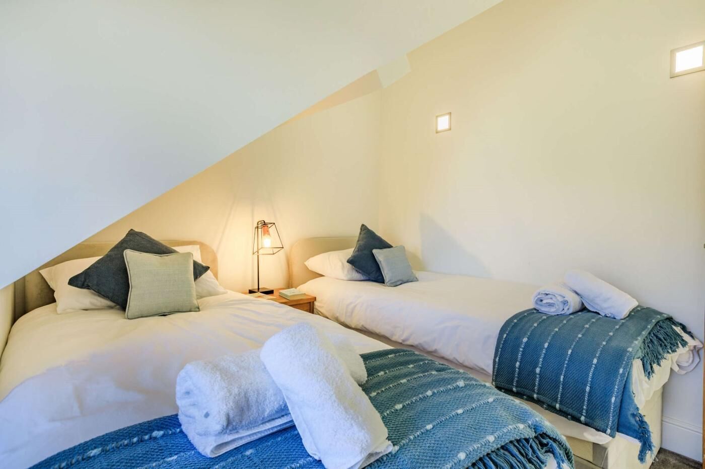 A room with two beds, a nightstand with a lamp, and a blue blanket on one bed.
