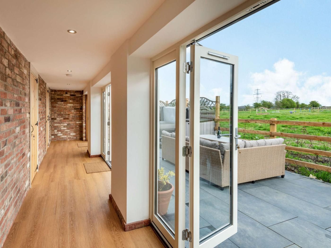 Un couloir avec un mur en briques, un plancher en bois et une porte coulissante en verre donnant sur un espace extérieur aménagé avec vue sur un champ verdoyant.