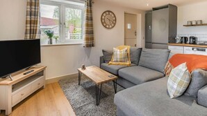 Ett vardagsrum med en platt-TV, en grå soffa, ett soffbord i trä och en köksdel med mikrovågsugn och kaffebryggare.