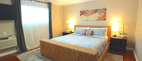 A bedroom with a bed, a ceiling fan, a nightstand with a lamp, and a wall-mounted air conditioner.