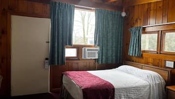 A room with a bed, a window with curtains, a thermostat, and wooden walls.