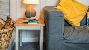 A cozy living area with a grey sofa, a yellow pillow, a small wooden side table with a lamp and books, and a wicker basket. - 1 Bed in Wadebridge (oc-87740) (Wadebridge)