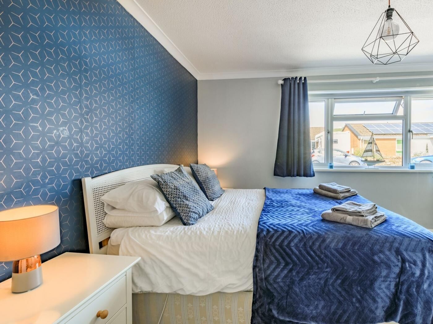 Et soveværelse med en seng, et natbord, en lampe, et vindue med gardiner og en hængende lysarmatur.