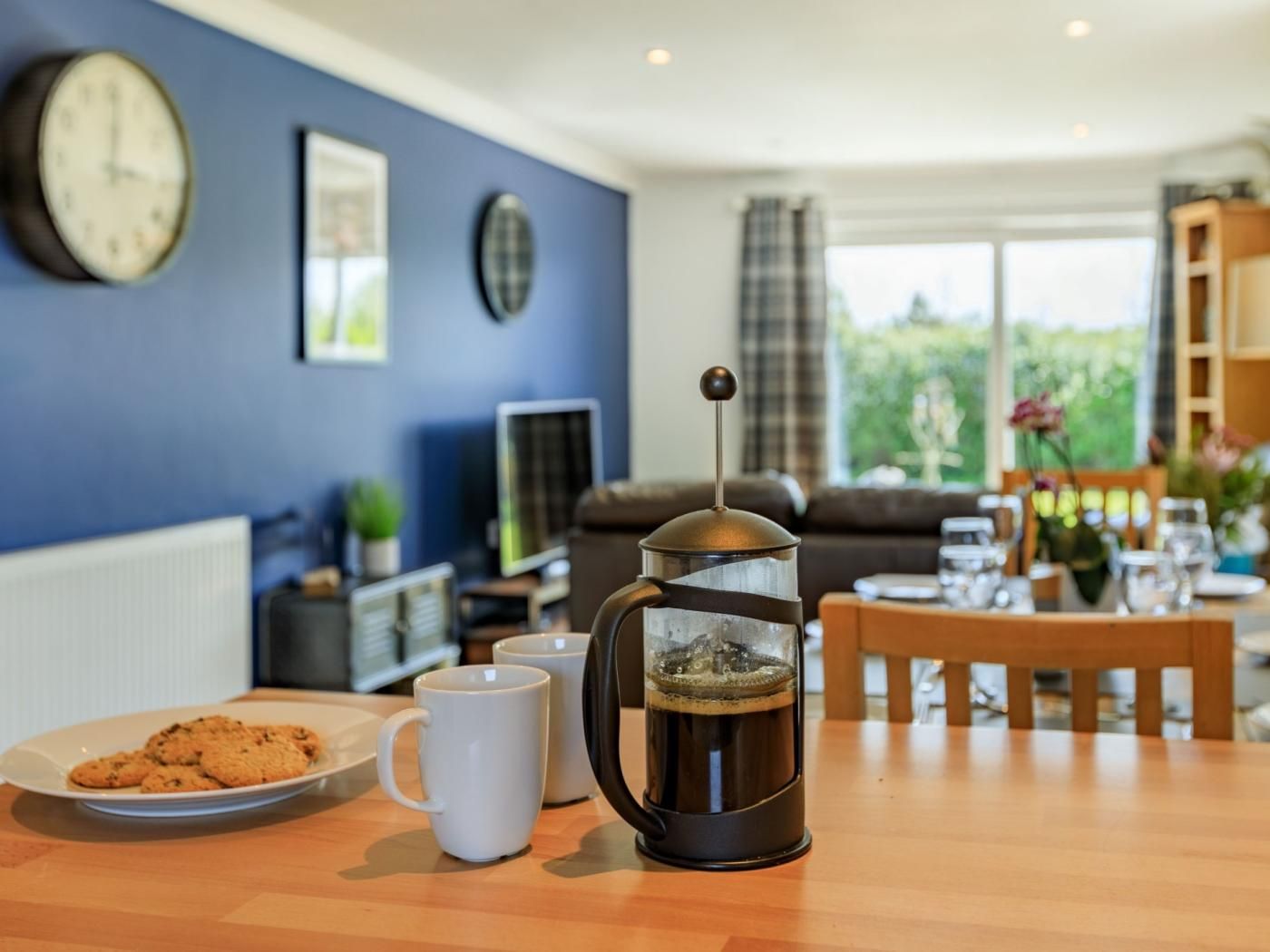 A coffee maker on a table with a plate of cookies and two white mugs.