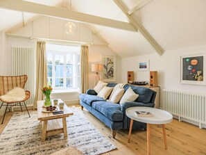 Ein gemütliches Wohnzimmer mit einem blauen Sofa, einem Holztisch, einem Korbstuhl und Blick ins Freie durch ein großes Fenster.