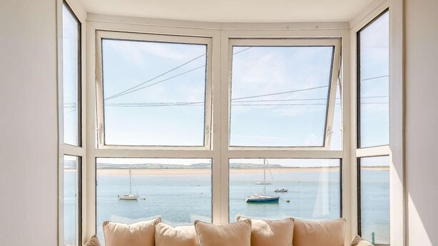 Un ventanal con vista al mar y barcos, un sofá escocés con cojines, y una pared blanca.
