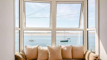 A bay window with a view of the sea and boats, a plaid sofa with cushions, and a white wall.